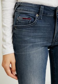 Tmavě modré džíny slim fit, s malou přední kapsou a červenými a bílými akcenty. Zapínání na knoflík a zip v pase.