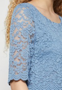 Bluza iz modrega cvetličnega čipkastega materiala s polovičnimi rokavi, ki jo nosi oseba s kodrastimi blond lasmi, ki razkriva detajle ovratnika in ramen.