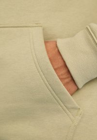 Main partiellement dans la poche avant d'un sweat-shirt beige clair avec des coutures détaillées et un poignet en côte visible.
