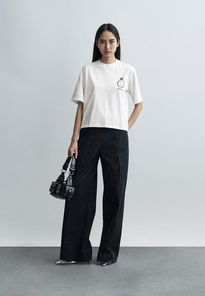 Κοντομάνικο λευκό τοπ με τύπωμα και μαύρο παντελόνι με φαρδιές μπατζάκια. Μαύρη τσάντα με υφή και ασημένιες λεπτομέρειες. Ασημένια μυτερά παπούτσια.