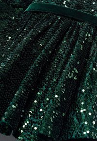 Tessuto verde con paillettes, caratterizzato da una superficie testurizzata e da paillettes circolari che riflettono la luce. La bordatura in velluto offre un contrasto in vita.