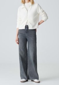 Hvid fleece zip-up jakke, hvid højhalset top, brede grå denim jeans og grå sneakers med hvide accenter. Casual outfit med neutrale farver.