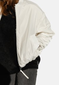 Weiße Bomberjacke mit glänzender Satin-Oberfläche, geribbeltem Kragen und elastischen Bündchen. Detail: silberner Reißverschluss und seitliche Taschen.