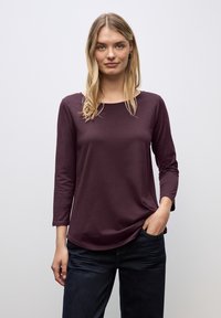 Bordeaux rood langemouwen top gemaakt van zachte stof met een ronde halslijn, een ontspannen pasvorm en een lichte kromming aan de onderkant, gecombineerd met donkere jeans.