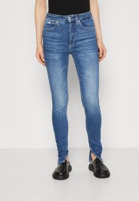 High-waisted blue denim skinny jeans with a smooth texture, two front pockets, and split hems. Paired with black slip-on shoes.