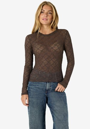 Femme aux longs cheveux blonds portant un haut en dentelle florale marron transparente à manches longues et un jean bleu, debout devant un fond uni.