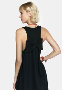 Czarna sukienka bez rękawów z odkrytymi plecami, z dużą kokardą na górze pleców. Miękka, lekko opływająca tkanina z luźnym krojem.