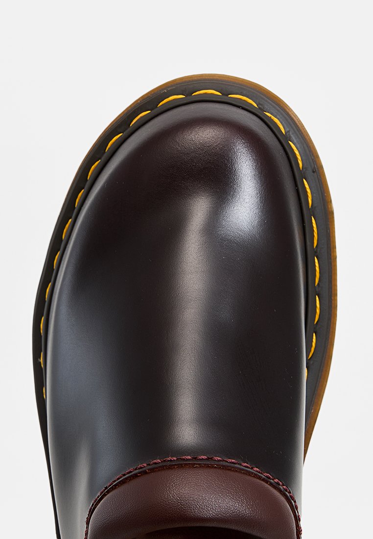 Zapato de cuero marrón con puntera redondeada, que presenta costuras amarillas en una suela negra. Textura suave con un acento burdeos en contraste cerca del talón.