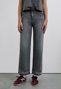 Vaqueros de pierna ancha en gris con lavado descolorido y pequeños tachuelas; combinados con zapatillas burdeos y un cinturón a juego. Tela de algodón, diseño casual.