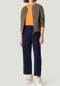 Veste matelassée olive, chemise orange, pantalon large bleu foncé, et baskets beige clair, avec une fermeture éclair à l'avant et des poignets côtelés.