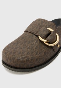 Brown patterned slip-on shoe with a textured surface, featuring a wide strap and a gold circular buckle accent. Rubber sole.