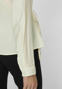 Blouse plissée jaune clair à manches longues, avec des détails de poignets à boutons et des liens sur les côtés. Portée avec un pantalon noir, présentée sur fond neutre.