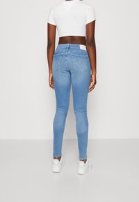 Jeans skinny azul claro con una cintura de media altura, bolsillos traseros y un parche de cuero blanco en la cinturilla, combinados con zapatillas blancas.