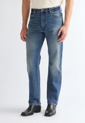 Homme portant un jean bleu délavé droit et des bottines noires à talons, debout devant un fond blanc uni.