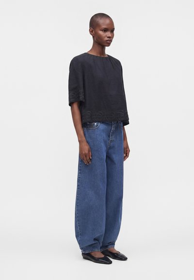 Femme aux cheveux courts portant un haut noir ample à manches courtes, un jean bleu large et des chaussures plates noires, debout devant un fond uni.