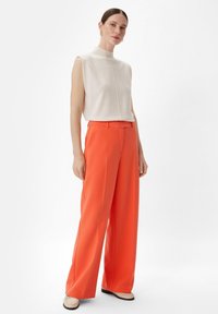 Beżowy top bez rękawów o wysokim kołnierzu i centralnym szwie, zestawiony z szerokimi spodniami w jaskrawym pomarańczowym kolorze, wykonanymi z gładkiego materiału.