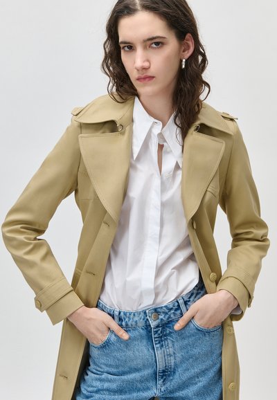 Γυναίκα που φοράει μπεζ παλτό τρανσέ, λευκό πουκάμισο με κουμπιά, ανοιχτό μπλε τζιν, με τα χέρια στις τσέπες, στέκεται μπροστά σε ένα απλό ανοιχτόχρωμο φόντο.