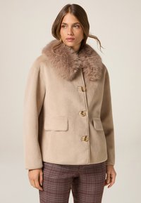 Abrigo de lana beige con un lujoso cuello de piel marrón, cuatro botones y dos bolsillos laterales. Presenta una forma cuadrada y una textura suave.