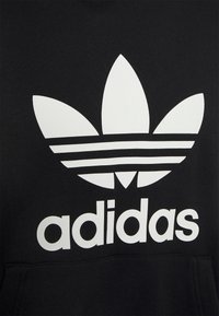 Fekete kapucnis felső nagy fehér Adidas logóval, trefoil dizájnnal és három vízszintes csíkkal. Puha, textúrált anyagból készült.