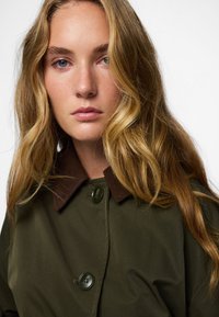 Oliivroheline mantel pruunist korterist kaelusega, millel on suured nööbid ja sile, tekstuurne kangas. Juuksed voolavad üle õlgade.