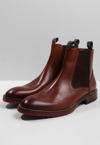 Bottes Chelsea en cuir marron avec panneaux latéraux élastiques, languettes de tirage, bouts arrondis, et une semelle en caoutchouc rouge contrastante. Finition lisse, design élégant.
