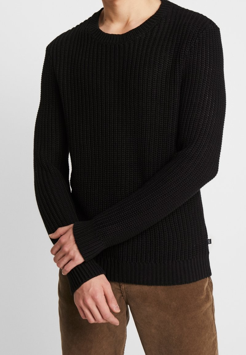 Personne portant un pull noir en maille à manches longues et un pantalon en velours côtelé marron, les mains doucement jointes devant.