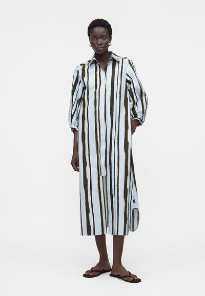 Γυναίκα που φοράει μακρύ, φαρδύ φόρεμα-πουκάμισο με κάθετες μαύρες και λευκές ρίγες, μανίκια τριών τετάρτων και σκούρα σανδάλια.