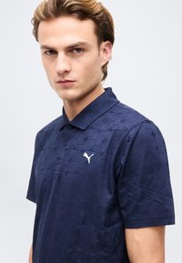 Jeune homme aux cheveux châtain clair portant un polo Puma bleu marine avec des motifs géométriques et un logo Puma blanc sur la poitrine.