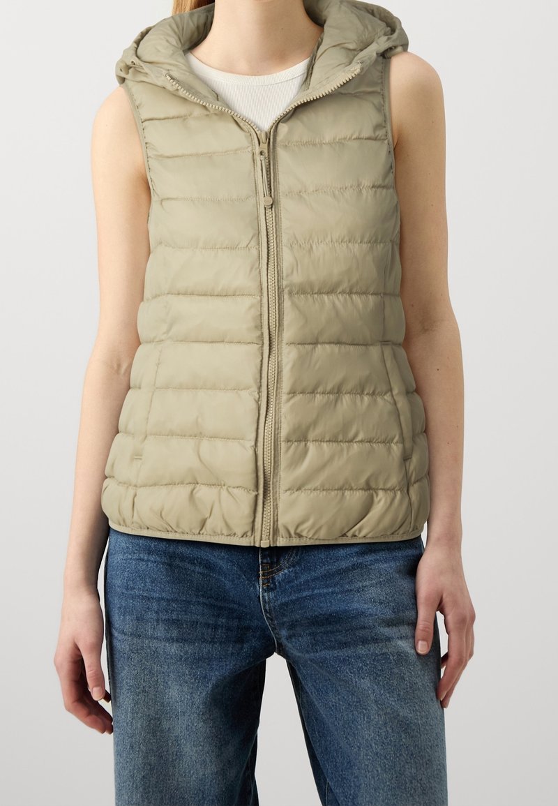 Veste matelassée sans manches beige à capuche zippée, portée sur un haut blanc avec un jean bleu sur une personne debout.