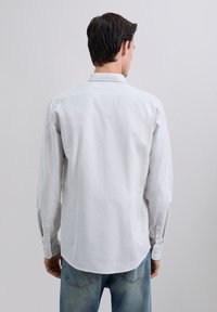 Uomo che indossa una camicia a righe grigio chiaro con maniche lunghe e jeans blu, in piedi di spalle contro uno sfondo bianco semplice.