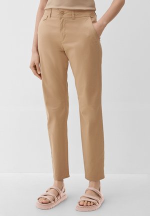 Beige bukser med lige ben og lommer foran og bagpå, båret med lyserøde sandaler med stropper på en lysegrå baggrund.