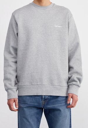Persona con sudadera Carhartt gris claro y vaqueros azules, de pie contra un fondo liso y claro, con las manos relajadas a los lados.