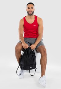 Red sleeveless tank top with a small white logo, gray shorts, and a black drawstring bag. White sneakers with textured sole detail.