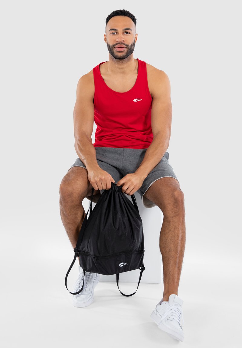 Red sleeveless tank top with a small white logo, gray shorts, and a black drawstring bag. White sneakers with textured sole detail.