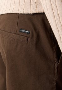 Beige cable knit sweater tucked into brown pants with a Scotch & Soda Amsterdam label above the back pocket.