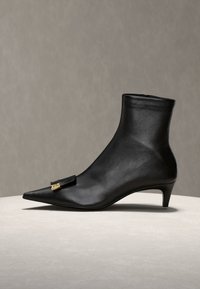 Bottine noire en cuir avec bout pointu, petit talon kitten et petite boucle dorée sur le devant sur fond neutre.