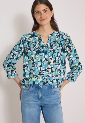 Femme aux cheveux foncés portant un chemisier bleu et blanc à fleurs et un jean bleu clair, debout les mains sur les hanches devant un fond uni.