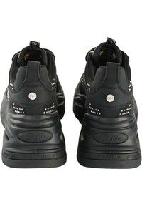 Fekete sportcipők struktúrált külsővel, tüskés díszítéssel a sarkakon, és vastag gumitalppal, amelyen kör alakú logó és húzófülek találhatóak.