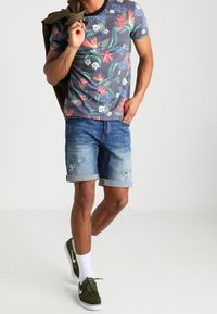Hombre luciendo una camiseta de estampado floral, pantalones cortos de mezclilla desgastados y arremangados, calcetas blancas y zapatillas verdes, sosteniendo una chaqueta sobre el hombro.