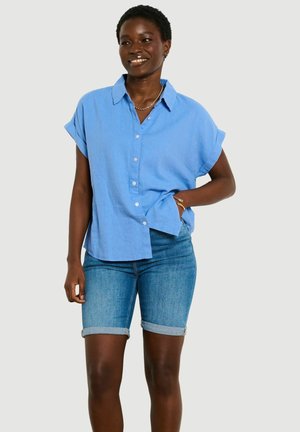 Usmievavá žena stojaca s rukou v vrecku, oblečená v svetlomodrú krátku košeľu na gombíky a zrolované rifľové šortky na jednoliatom pozadí.