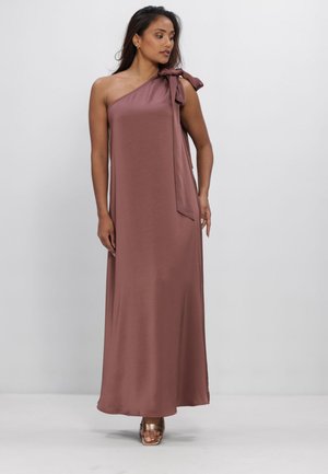 Vrouw draagt een mauve, asymmetrische maxi-jurk met een grote strik op de schouder, staand voor een effen lichtgrijze achtergrond.