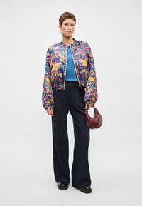 Veste bomber colorée avec des imprimés floraux et animaux, associée à un haut bleu et un pantalon large rayé. Tenant un sac à main bordeaux.