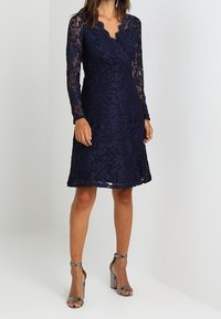 Námořnické krajkové šaty s dlouhými rukávy a výstřihem ve tvaru vlnky, s přiléhavým korzetem a rozšířenou sukní, doplněné o metalické sandály na vysokém podpatku.