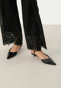 Juodos smailianosiai slingback bateliai su žemu kulnu, pagaminti iš lygaus odos. Dėvimi su juodais kelnėmis, puoštomis nėriniu, turinčiomis tekstūrinį raštą.