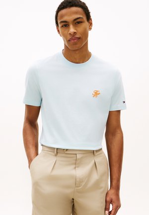 Junger Mann trägt ein hellblaues T-Shirt mit orangefarbenem Logo auf der Brust und beigefarbene Hosen, Hände in den Taschen, vor einem schlichten weißen Hintergrund.