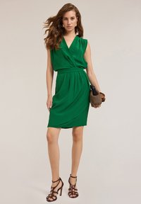Grünes ärmelloses Kleid mit V-Ausschnitt, geraffter Taille und drapiertem Saum. Glattes Material und knielange Ausführung. Trägt dazu Riemchen-Absätze.