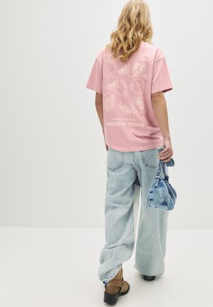 Person wearing a pink T-shirt with pomegranate drawings and text, light blue wide-leg jeans, brown boots, and holding a small denim bag.