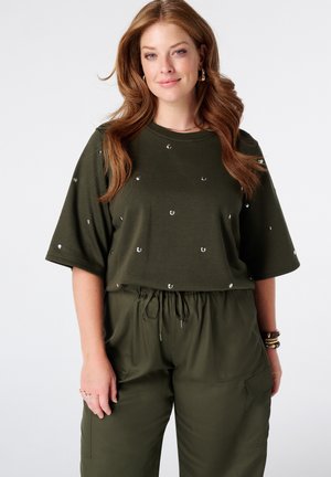 Femme aux longs cheveux roux portant un haut manches courtes vert olive avec de petits clous métalliques et un pantalon à cordon assorti.