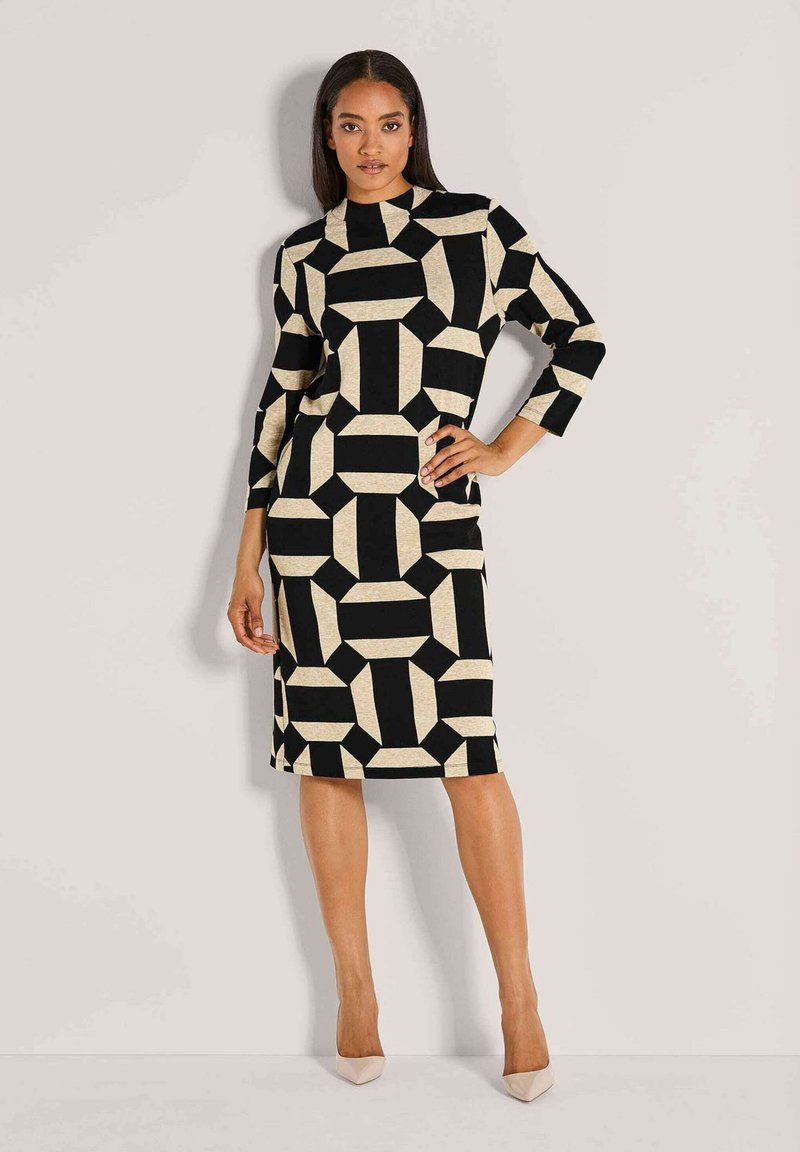 Frau steht mit einer Hand auf der Hüfte, trägt ein knielanges schwarzes Kleid mit beige-farbenen geometrischen Mustern und beige-farbenen Absätzen vor einem einfarbigen Hintergrund.