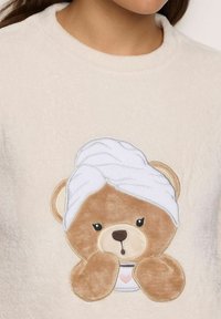 Orsetto di peluche con un asciugamano bianco avvolto sulla testa che tiene una tazza con un cuore rosa su un morbido tessuto color crema.